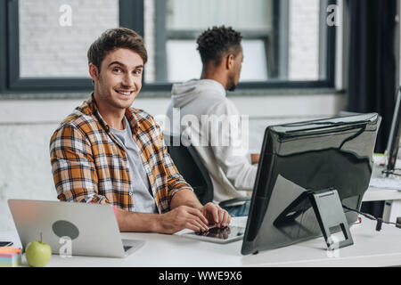 cheerful programmer smiling at camera while sitting near african american programmer Stock Photo