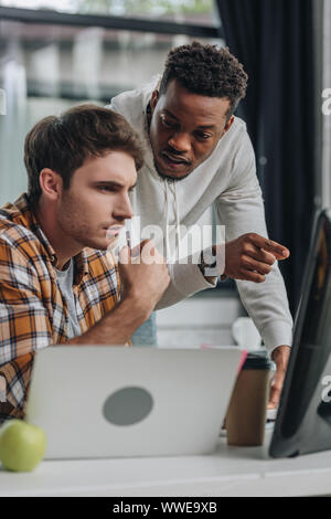 serious african american programmer pointing at computer monitor near thoughtful colleague Stock Photo