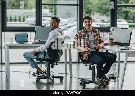 young multicultural programmers smiling at camera while sitting in office chairs Stock Photo