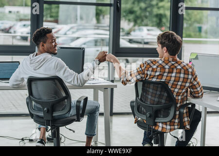 young multicultural programmers doing fist bump while sitting in office together Stock Photo