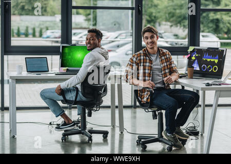 two young multicultural programmers looking at camera while sitting near computer monitors with graphs and charts on screen Stock Photo