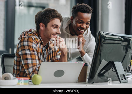 two cheerful multicultural programmers looking at computer monitor while working in office together Stock Photo