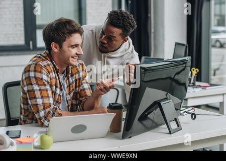 two smiling multicultural programmers pointing at computer monitor while working in office together Stock Photo