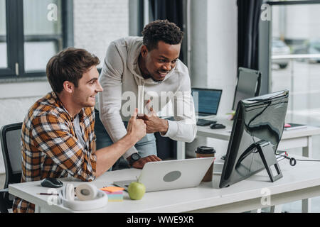 two cheerful multicultural programmers showing thumbs up while looking at computer monitor Stock Photo