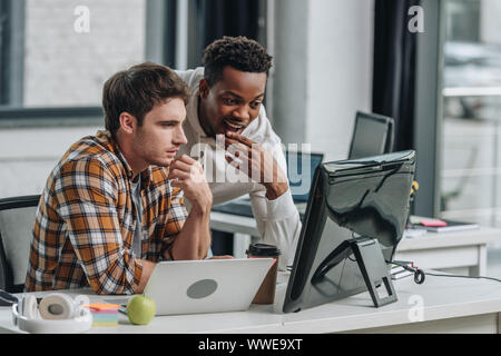 two thoughtful multicultural programmers looking at computer monitor while working in office together Stock Photo