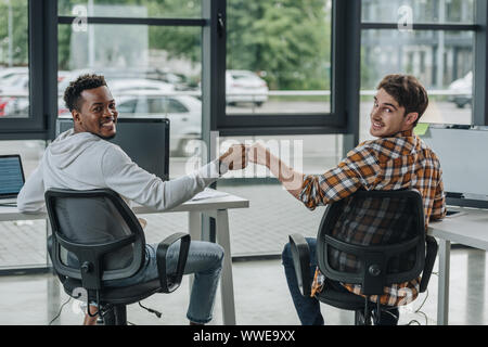two cheerful multicultural programmers looking at camera while doing fist bump Stock Photo