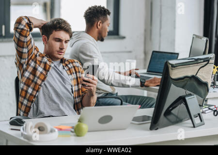 thoughtful programmer using smartphone while sitting at workplace near african american colleague Stock Photo