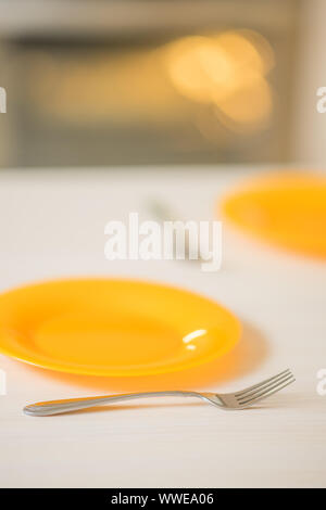 served table with orange plates and forks, waiting for food from the oven in the background. Stock Photo