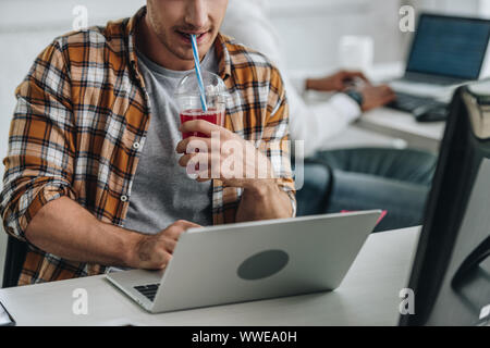 partial view of young programmer drinking juice while working on laptop near african american colleague Stock Photo