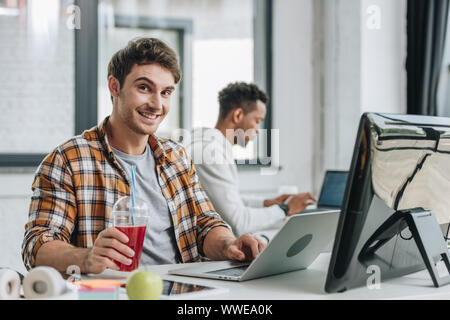 cheerful programmer looking at camera while sitting near african american colleague Stock Photo