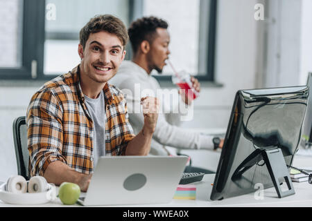 happy programmer looking at camera and showing winner gesture while sitting near african american colleague Stock Photo