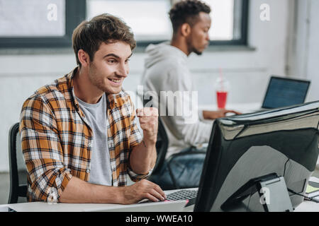 cheerful programmer showing winner gesture while working near african american colleague Stock Photo