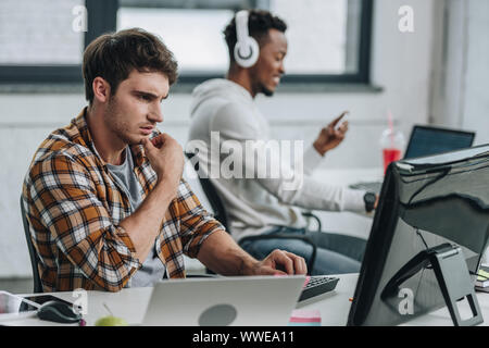 selective focus of thoughtful programmer working near african american colleague in office Stock Photo
