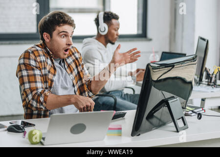 selective focus of shocked programmer gesturing while working near african american colleague Stock Photo