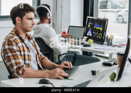back view of african american programmer in headphones sitting near computer monitor with graphs and charts on screen near colleague in office Stock Photo
