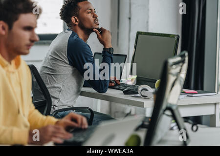 selective focus of thoughtful african american programmer looking away while sitting near colleague in office Stock Photo