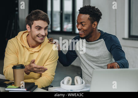 happy young programmer pointing at computer monitor while sitting near african american colleague in office Stock Photo