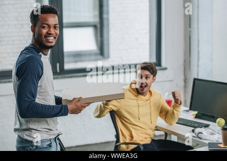cheerful african american programmer holding pizza box while colleague showing yes gesture Stock Photo