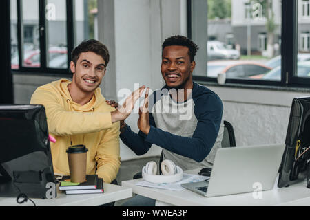 two multicultural programmers giving high five and smiling at camera in office Stock Photo