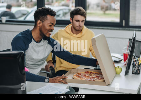 smiling african american programmer opening pizza box while sitting near colleague in office Stock Photo