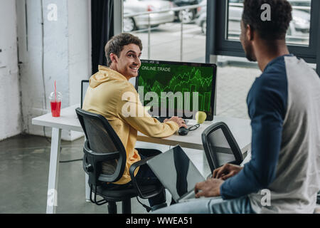 cheerful programmer sitting at computer monitor with online trade on screen and looking at african american colleague Stock Photo