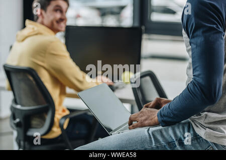 partial view of african american programmer sitting on desk with laptop near smiling colleague Stock Photo