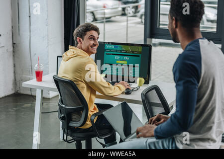 KYIV, UKRAINE - JULY 29, 2019: smiling programmer using computer with Sportsbet website on screen while his african american colleague sitting on desk Stock Photo