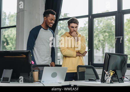 smiling african american programmer standing near thoughtful colleague in office Stock Photo