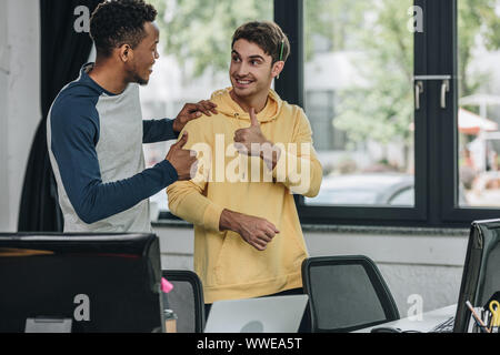 cheerful multicultural programmers looking at each other and showing thumbs up Stock Photo