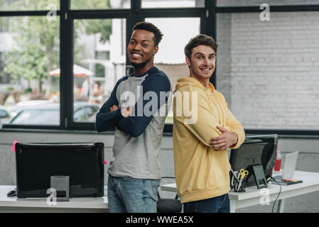 two happy multicultural programmers standing back to back with crossed arms and smiling at camera in office Stock Photo