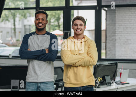 two cheerful multicultural programmers standing with crossed arms and smiling at camera in office Stock Photo