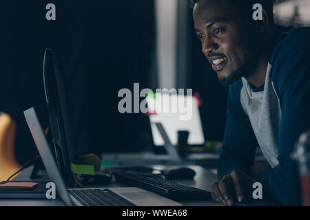 young african american programmer smiling while looking at computer monitor at night in office Stock Photo