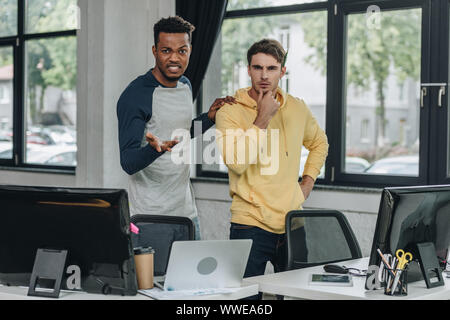 irritated african american programmer gesturing while standing near colleague in office Stock Photo