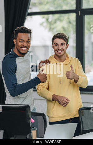 two happy multicultural programmer showing thumbs up while smiling at camera in office Stock Photo