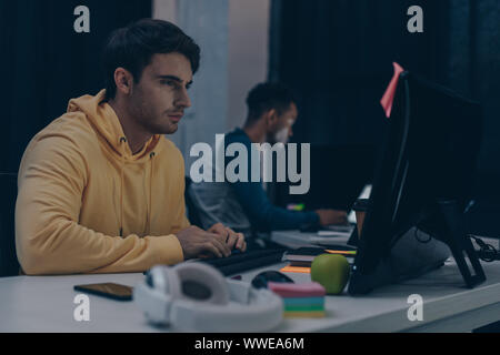 selective focus of serious programmer looking at monitor while working at night near african american friend Stock Photo