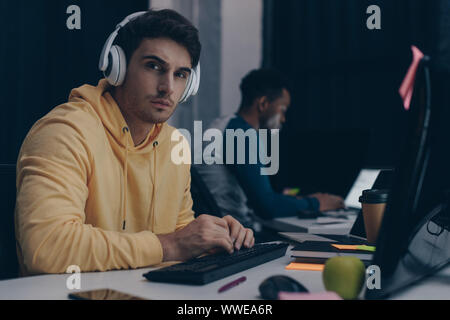 selective focus of young programmer in headphones looking at camera while working near african american colleague Stock Photo