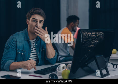 selective focus of shocked programmer looking at camera while working near african american colleague Stock Photo