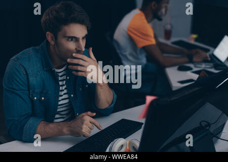selective focus of shocked programmer working at night in office near multicultural friend Stock Photo