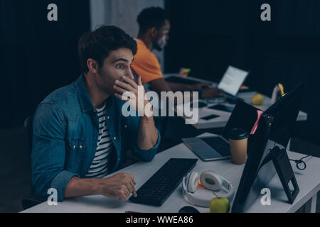 selective focus of surprised programmer looking at monitor while working near african american colleague Stock Photo
