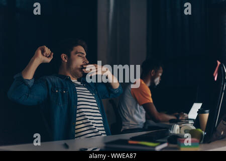 selective focus of tired programmer yawning while sitting near african american colleague and night in office Stock Photo