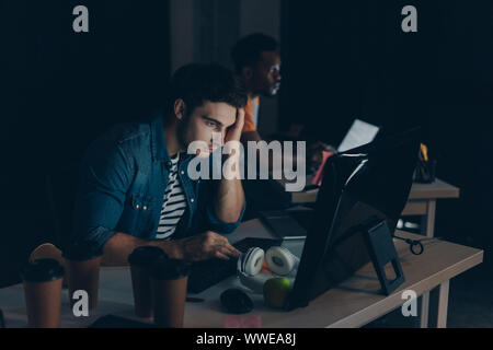 selective focus of thoughtful programmer working at night in office near african american colleague Stock Photo