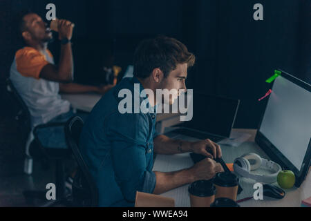 selective focus of attentive programmer working on computer near african american colleague drinking coffee to go Stock Photo
