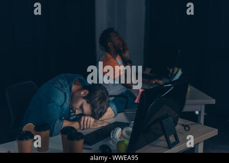 exhausted programmer sleeping at workplace while african colleague working at night in office Stock Photo