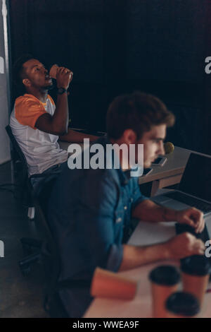 selective focus of african american programmer drinking coffee to go while working near multicultural colleague at night in office Stock Photo