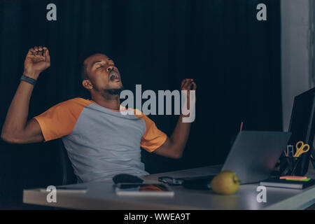 tired african american programmer yawning while sitting at night in office Stock Photo