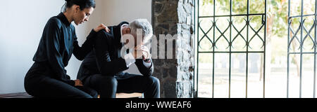 panoramic shot of attractive woman sitting near upset senior man in cemetery Stock Photo