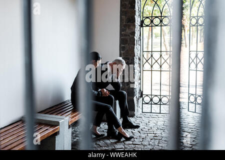 beautiful woman sitting on bench near sad senior man in cemetery Stock Photo