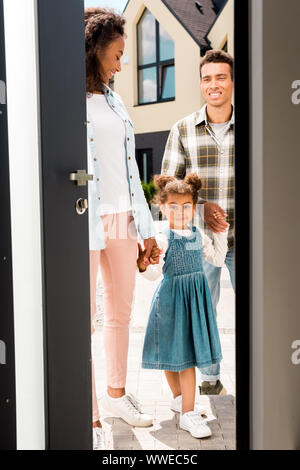 full length view of kid looking at camera while holding hands with african american parents and walking into house Stock Photo