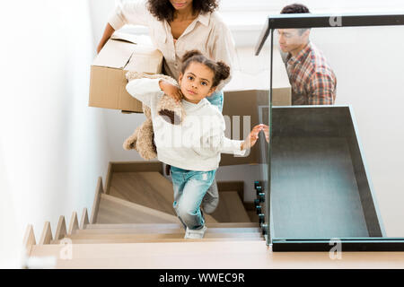 cropped view of african american woman going upstairs with box while daughter holding toy and father walking after mother Stock Photo