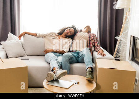 full length view of husband sleeping on sofa while wife sitting near man and looking at camera Stock Photo